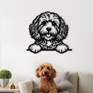 Può includere: Arte da parete in metallo nero di un cane che sporge da una superficie. Il cane ha i capelli ricci e guarda direttamente la telecamera. Un cane marrone è seduto su un divano bianco sullo sfondo.