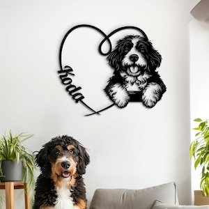 May include: A black metal wall art piece featuring a dog inside a heart shape, with the name "Koda" in cursive. A Bernese Mountain Dog puppy sits on a gray beanbag chair. The dog has black, brown, and white fur.