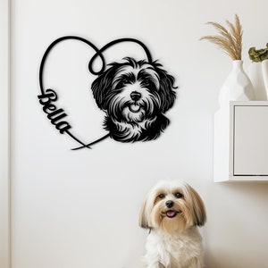 Op de afbeelding: Een zwart metalen hartvormige silhouet met de naam "Bella" erin gegraveerd. Een zwart-witte hondenkop bevindt zich in het hart. Een witte hond zit op een beige hondenbed voor de wandkunst.