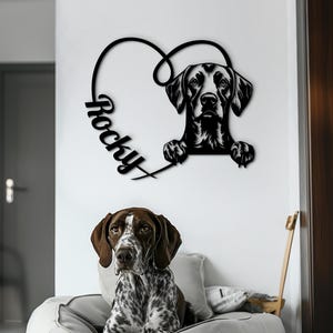 May include: Black metal wall art of a dog's head in a heart shape, with the name "Rocky". A real dog with brown and white spots is lying on a round, light grey dog bed. The dog is a German Shorthaired Pointer.