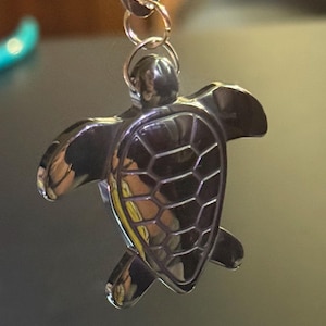 May include: A polished silver turtle keyring. The turtle charm features a detailed shell pattern and is connected to a silver chain and ring. This keyring is a decorative accessory.