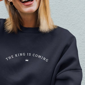 Puede incluir: Una mujer lleva una sudadera negra con el texto "THE KING IS COMING" y un gr&aacute;fico de corona impreso en la parte delantera. Est&aacute; sonriendo y haciendo un signo de paz con la mano.