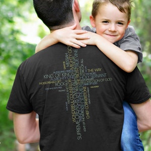 May include: A black t-shirt with a large white cross made of words related to Jesus Christ. The words are in yellow and green. A man is holding a young boy in his arms. The boy is smiling and looking at the camera.