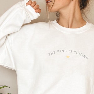 Puede incluir: Sudadera blanca con el texto "THE KING IS COMING" en letras grises y un gr&aacute;fico de corona dorada.