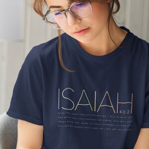 May include: A navy blue t-shirt with the word "ISAIAH" printed in a multi-colored font. The text "43:2" is printed in a smaller font below the word "ISAIAH". The t-shirt has a quote from the Bible printed on it: "When you pass through the waters, I will be with you; and when you pass through the rivers, they will not sweep over you. When you walk through the fire, you will not be burned; the flames will not set you ablaze."