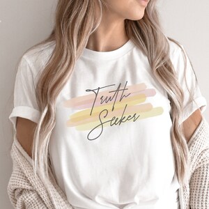 May include: White t-shirt with a graphic design of the words "Truth Seeker" in black script with a watercolor-style background of pink, orange, and yellow.