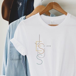 May include: White t-shirt with a graphic design of the word "Jesus" in colorful lines and the text "is Lord" below it.