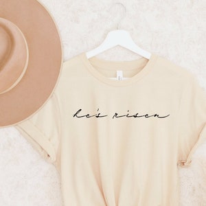 以下が含まれることがあります： 黒字で「He's Risen」と書かれたクリーム色のTシャツ。白いハンガーにかけられており、背景にはベージュの帽子があります。Tシャツはウエストで結ばれています。