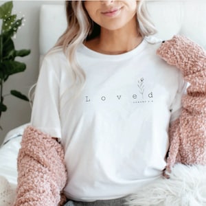May include: White t-shirt with the word "Loved" and a small floral design. The text "Romans 5:8" is written below the design.