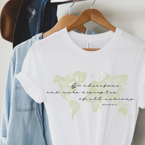 Op de afbeelding: Wit T-shirt met een groene wereldkaart afbeelding en de tekst "Ga daarom en maak discipelen van alle volken" met de bijbelverwijzing "Matthe&uuml;s 28:19".