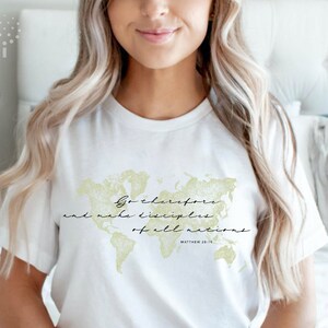 Op de afbeelding: Wit T-shirt met een gouden omtrek van een wereldkaart en de tekst "Ga daarom heen en maak alle volken tot mijn discipelen. Matthe&uuml;s 28:19"