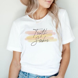 May include: White t-shirt with the text "Truth Seeker" in black script. The text is layered over a design of horizontal, abstract watercolor brushstrokes in shades of pink, orange, and yellow.