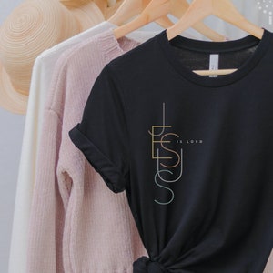 May include: Black t-shirt with a vertical graphic design of the word "Jesus" in colorful lines. The text "is Lord" is written in smaller font below the graphic.