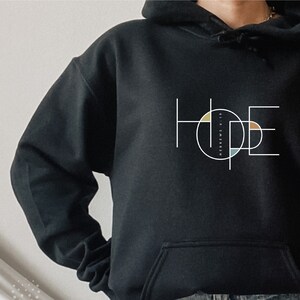 May include: Black hoodie with a white graphic of the word "HOPE" in a geometric design with colourful circles. The text "HEBREWS 10:23" is inside one of the circles.