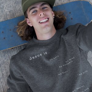 May include: A dark gray sweatshirt with the text "Jesus is..." printed on the front. The text is followed by a list of words: "my Lord", "my King", "my Strength", "my Hope", "my Life", "my Savior", and "my God".
