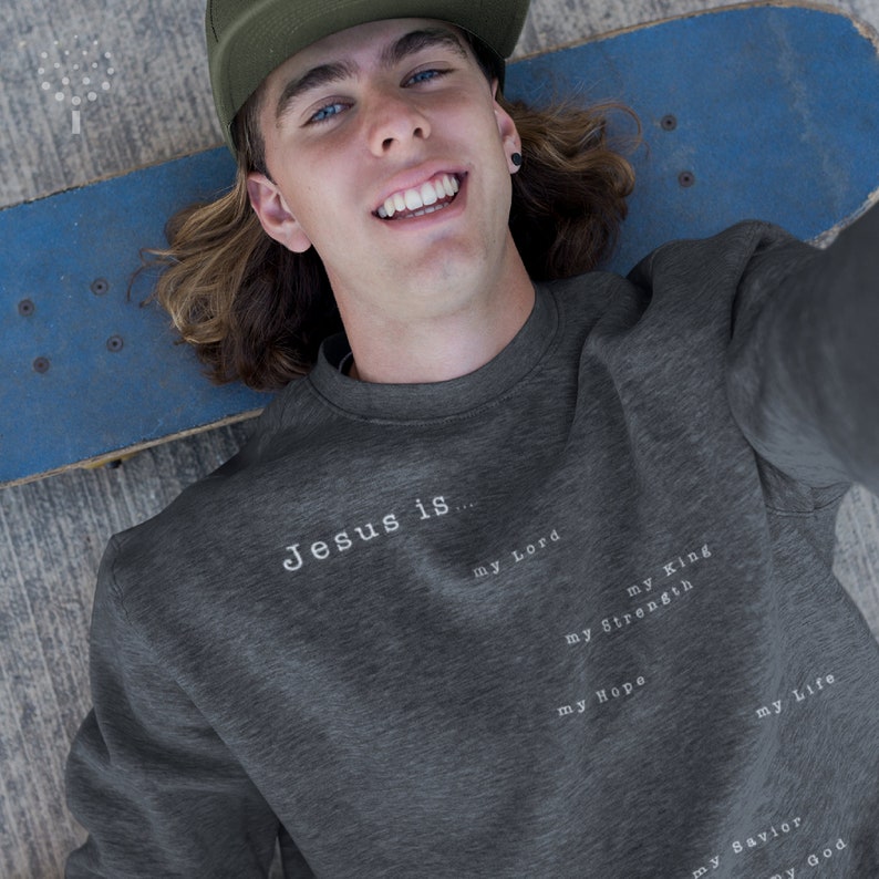 May include: A dark gray sweatshirt with the text "Jesus is..." printed on the front. The text is followed by a list of words: "my Lord", "my King", "my Strength", "my Hope", "my Life", "my Savior", and "my God".