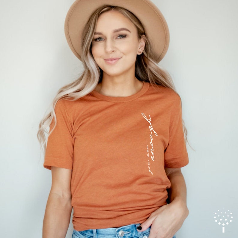 May include: A rust-colored t-shirt with the text "you are enough" written vertically in white on the right side of the shirt.