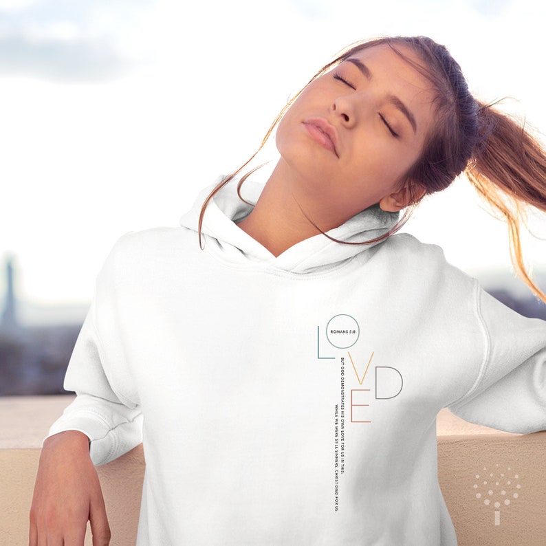 May include: White hoodie with a graphic design that says "LOVED" with a vertical line and the text "ROMANS 5:8 But God demonstrates his own love for us in this: While we were still sinners, Christ died for us."