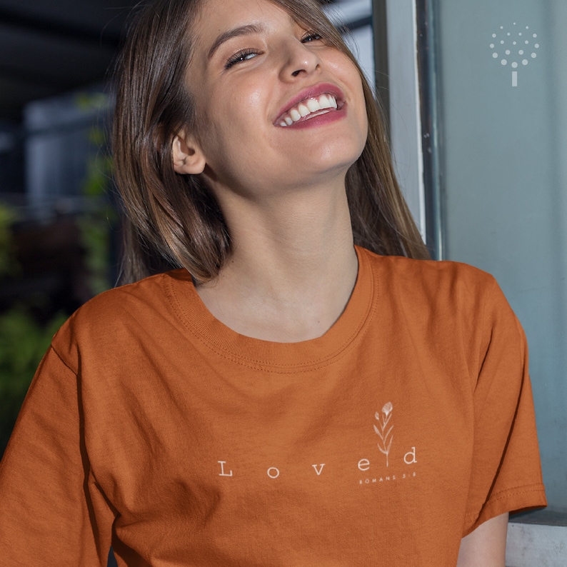 May include: A woman wearing a rust-colored t-shirt with the word "Loved" and a small floral design. The text "Romans 5:8" is written in smaller font below the floral design.