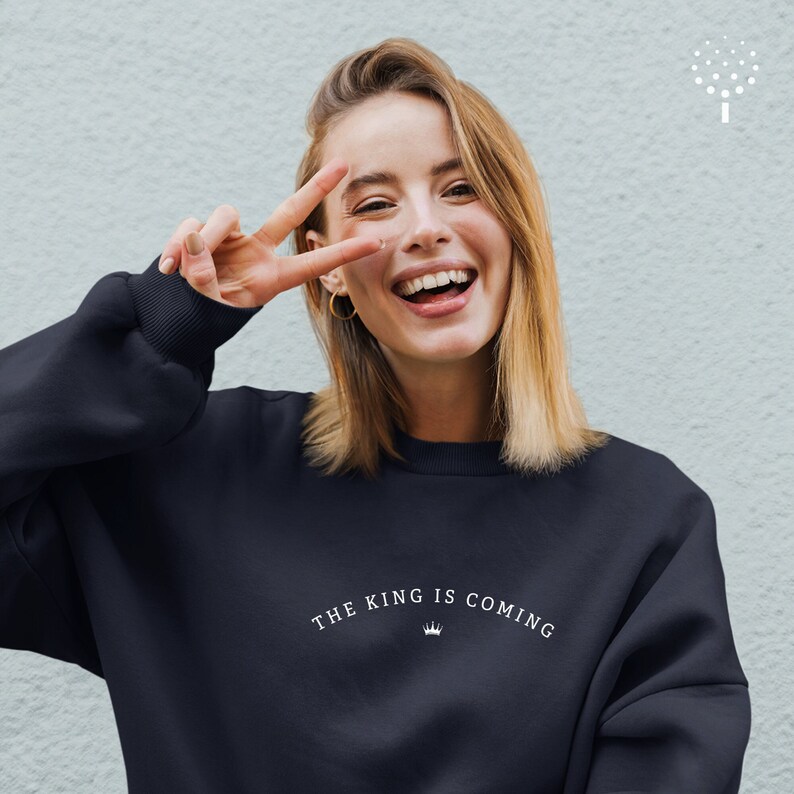 Puede incluir: Una mujer lleva una sudadera negra con el texto "THE KING IS COMING" y un gr&aacute;fico de corona impreso en la parte delantera. Est&aacute; sonriendo y haciendo un signo de paz con la mano.