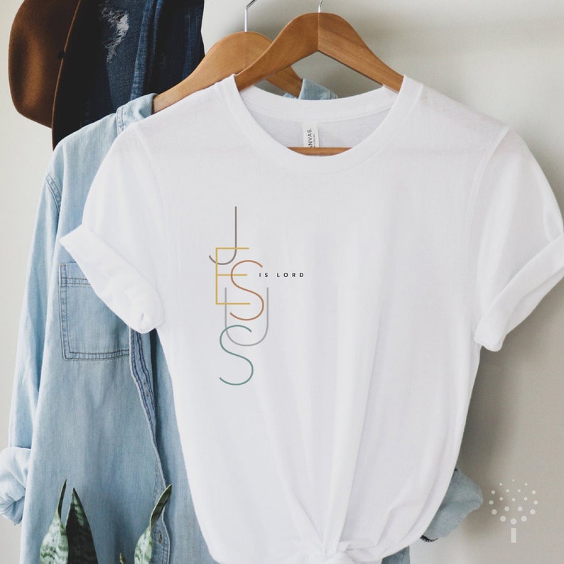 May include: White t-shirt with a graphic design of the word "Jesus" in colorful lines and the text "is Lord" below it.