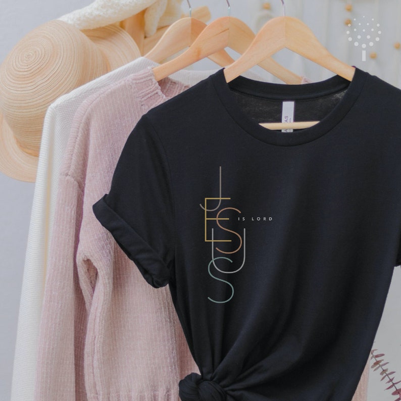 May include: Black t-shirt with a vertical graphic design of the word "Jesus" in colorful lines. The text "is Lord" is written in smaller font below the graphic.