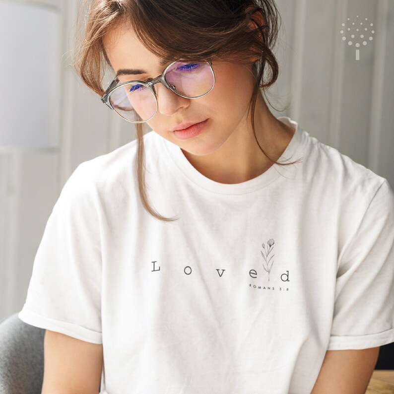 May include: White t-shirt with the word "Loved" and the text "Romans 5:8" printed on it. A small floral design is above the word "Loved".