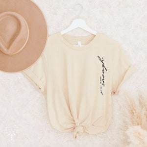 May include: A beige t-shirt with the text "you are enough" written in black on the left side of the shirt.