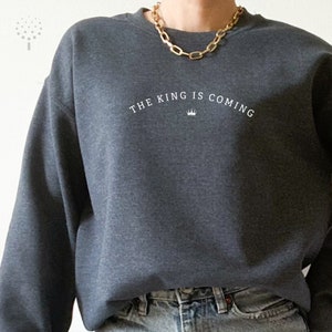 Puede incluir: Una sudadera gris oscuro con el texto "THE KING IS COMING" en letras blancas y un peque&ntilde;o s&iacute;mbolo de corona.