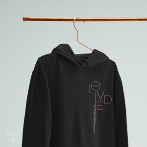May include: Black hoodie with a vertical graphic design that says "LOVED" in colorful letters. The graphic also includes the Bible verse "ROMANS 5:8" and the text "BUT GOD DEMONSTRATES HIS OWN LOVE FOR US IN THIS: WHILE WE WERE STILL SINNERS, CHRIST DIED FOR US."