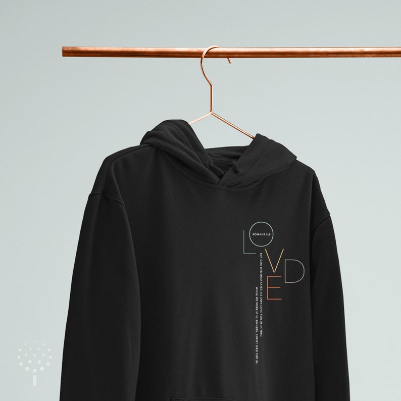 May include: Black hoodie with a vertical graphic design that says "LOVED" in colorful letters. The graphic also includes the Bible verse "ROMANS 5:8" and the text "BUT GOD DEMONSTRATES HIS OWN LOVE FOR US IN THIS: WHILE WE WERE STILL SINNERS, CHRIST DIED FOR US."
