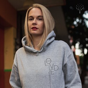 May include: A woman wearing a gray hoodie with the word "LOVED" printed vertically on the front. The word is surrounded by a circle with the text "SCHWANENK.K" inside.