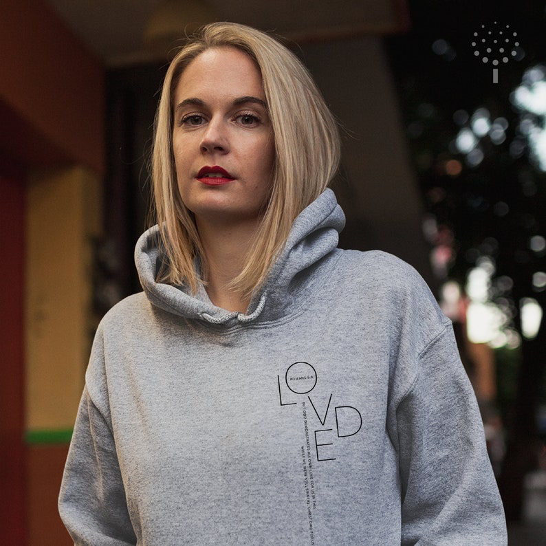 May include: A woman wearing a gray hoodie with the word "LOVED" printed vertically on the front. The word is surrounded by a circle with the text "SCHWANENK.K" inside.