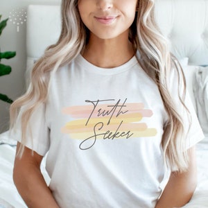 May include: White t-shirt with the text "Truth Seeker" in a handwritten font. The text is on a background of pastel pink, yellow, and orange watercolor stripes.