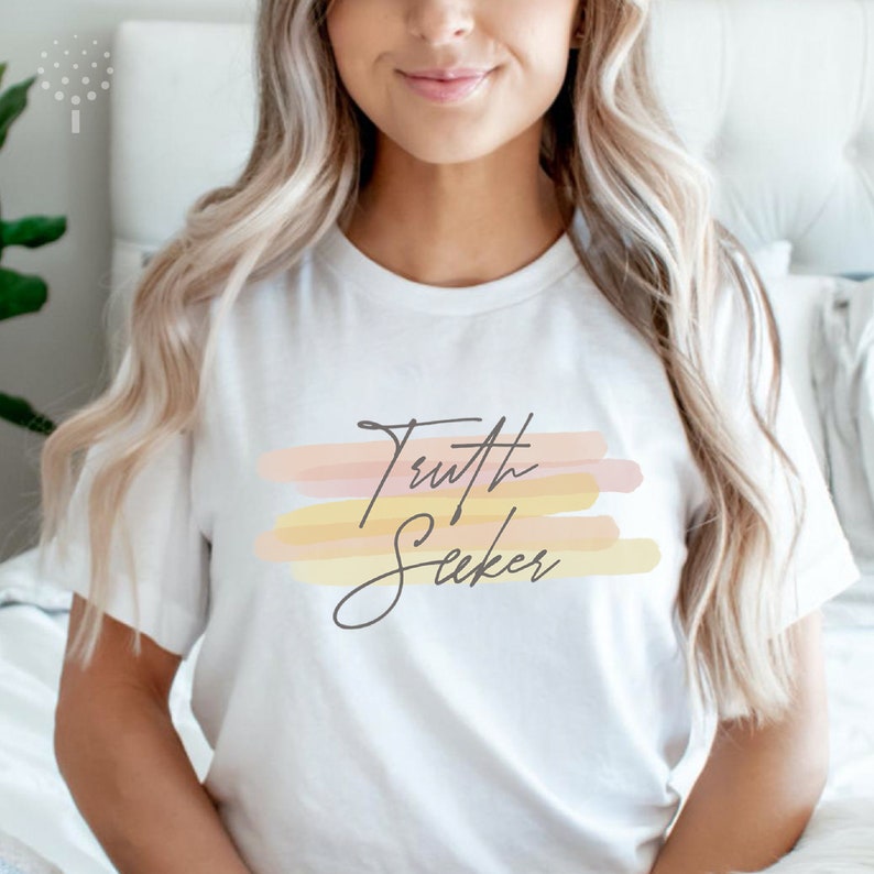 May include: White t-shirt with the text "Truth Seeker" in a handwritten font. The text is on a background of pastel pink, yellow, and orange watercolor stripes.