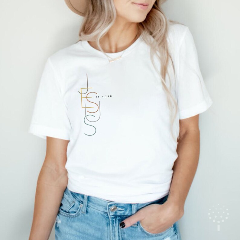May include: White t-shirt with a graphic design of the word "JESUS" in a vertical, stylized font. The word "IS LORD" is written in smaller font next to the "S" in "JESUS".