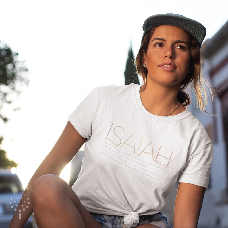 May include: A white t-shirt with the word "ISAIAH" printed in rainbow colors and the verse "For I am the Lord your God, who takes hold of your right hand and says to you, Do not fear; I will help you." Isaiah 41:13 printed in black below.
