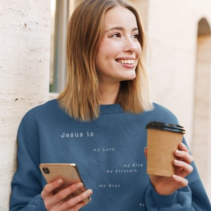 May include: A woman wearing a blue sweatshirt with the text "Jesus is... my Lord, my King, my Strength, my Hope" printed on it. She is holding a brown paper cup and a smartphone.