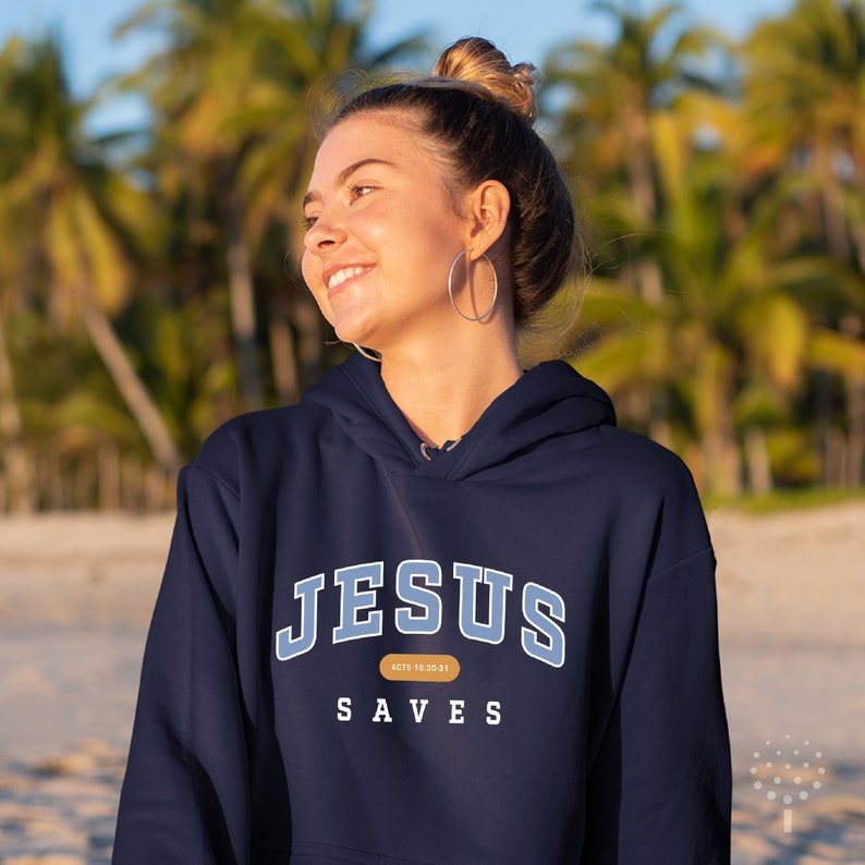 May include: A navy blue hoodie with the text "JESUS SAVES" in white and blue lettering. The text "ACTS 18:25-21" is in a small brown rectangle below the word "JESUS".