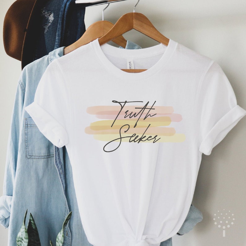 May include: White t-shirt with the text "Truth Seeker" in black script. The text is layered over three horizontal brushstrokes in shades of pink, yellow, and orange.