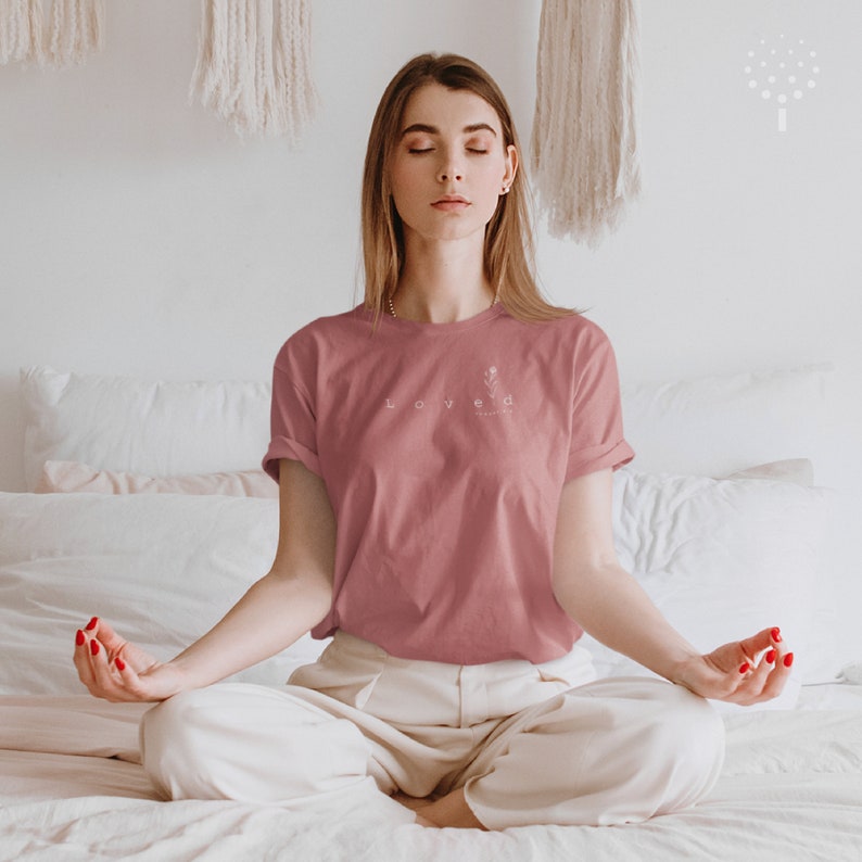 May include: A woman wearing a pink t-shirt with the word "Loved" and a floral design on the front. She is sitting in a meditative pose on a white bed with her eyes closed.