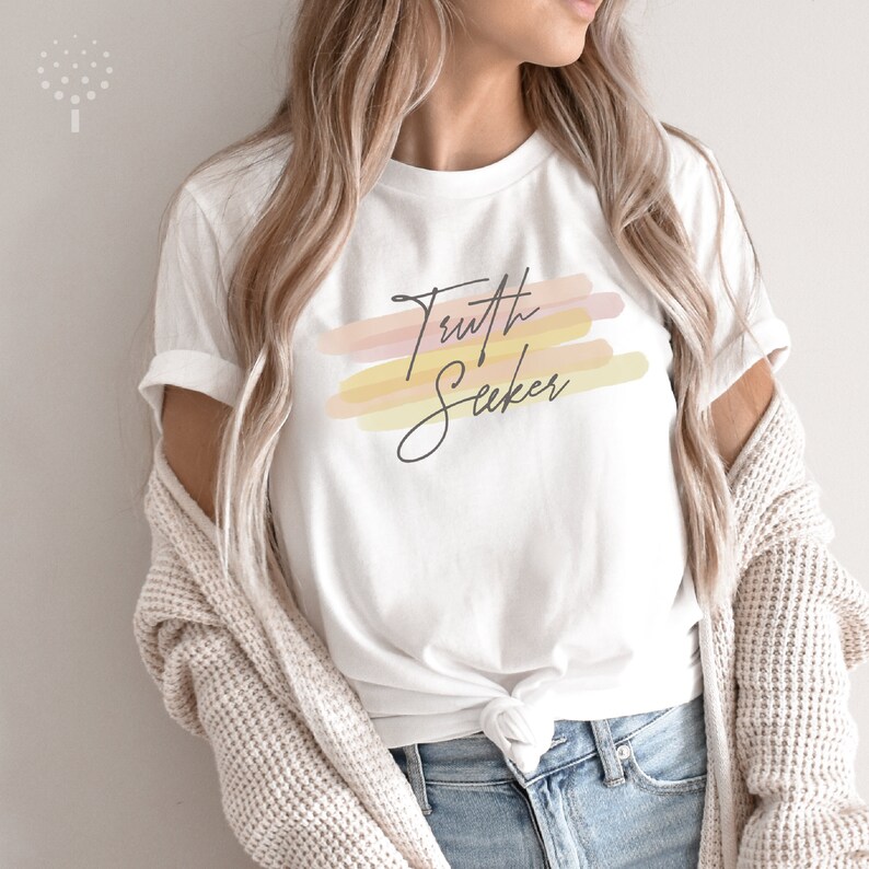 May include: White t-shirt with a graphic design of the words "Truth Seeker" in black script with a watercolor-style background of pink, orange, and yellow.