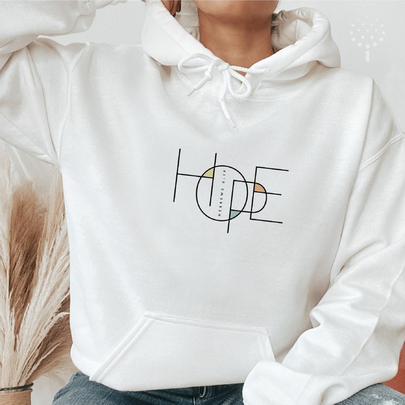 May include: White hoodie featuring a graphic design of the word "HOPE" in black outlined letters with colourful geometric shapes inside the letters. The text "Hebrews 6:19" is written inside the circle formed by the letters.