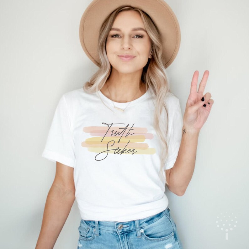 May include: A white t-shirt with the text "Truth Seeker" in black script. The text is layered over a design of horizontal watercolor stripes in shades of pink, orange, and yellow.