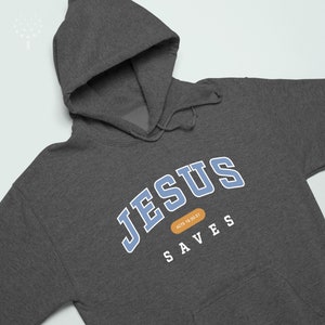 May include: A dark gray hooded sweatshirt with the text "JESUS SAVES" in blue and white lettering. The text "ACTS 18:30-31" is in a small orange rectangle.