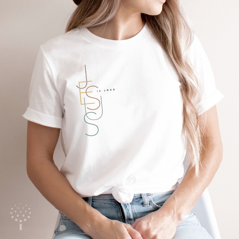 May include: White t-shirt with a graphic design of the word "JESUS" in a vertical, stylized font with the words "IS LORD" in smaller font above the "S".