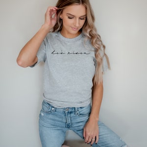以下が含まれることがあります： 「He's risen」の黒い文字が書かれたヘザーグレーのTシャツ。クルーネックのスタイルで、青いジーンズと合わせています。