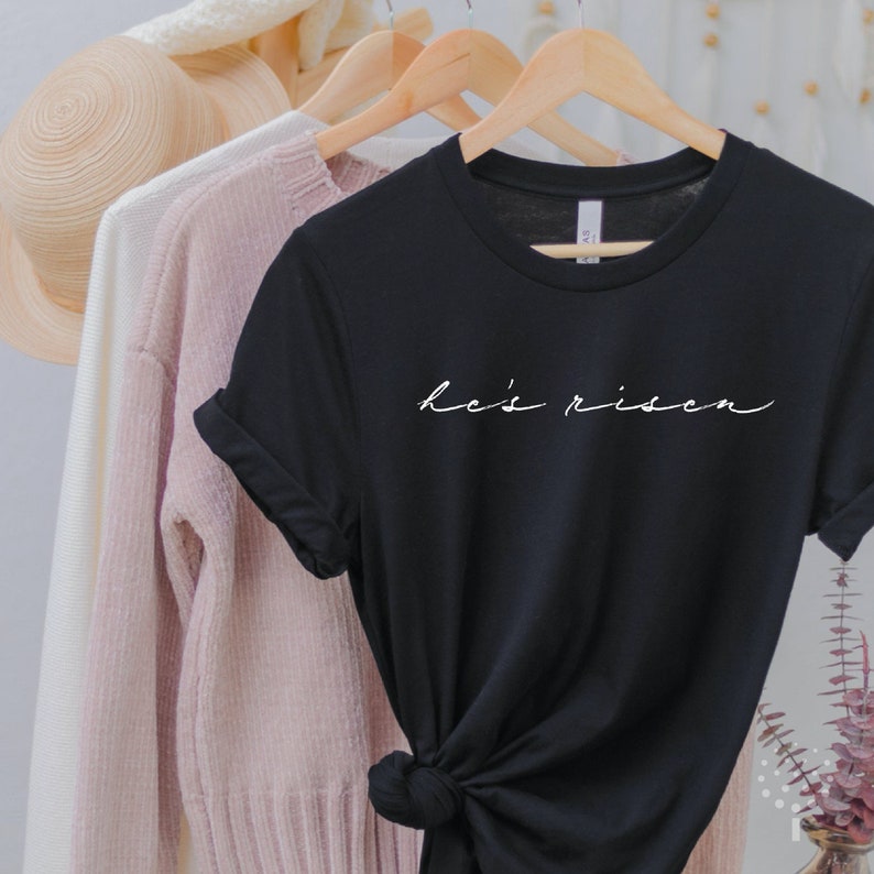 以下が含まれることがあります： 白い筆記体で「he's risen」と書かれた黒いTシャツ。ピンクのセーターと白い帽子など、他の衣類と一緒に木製のハンガーに掛けられています。