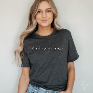 以下が含まれることがあります： 白い筆記体フォントで「He's risen」と書かれた濃い灰色のTシャツ。Tシャツは半袖でクルーネックです。モデルはブルージーンズを着用しています。