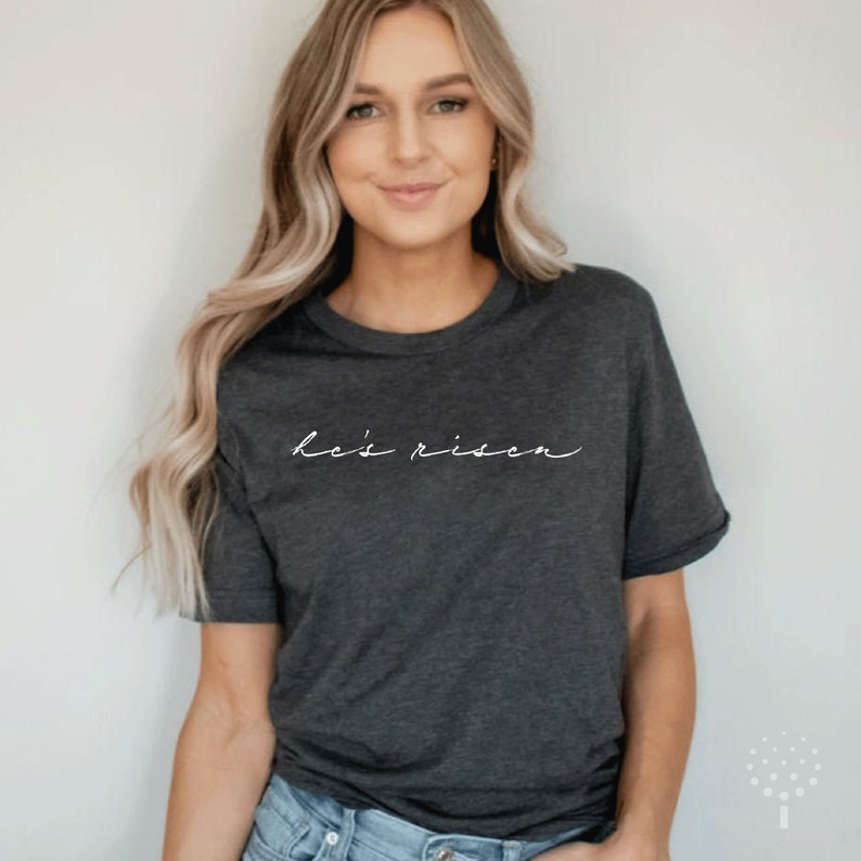 以下が含まれることがあります： 白い筆記体フォントで「He's risen」と書かれた濃い灰色のTシャツ。Tシャツは半袖でクルーネックです。モデルはブルージーンズを着用しています。